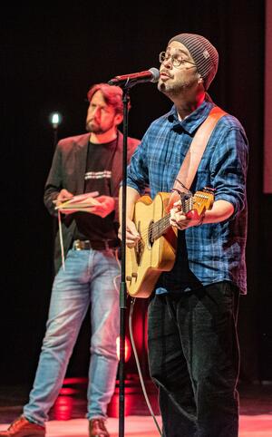 En mann spiller på gitar og synger med blårutete skjorte og lue en annen mann står i bakgrunnen og holder en bok